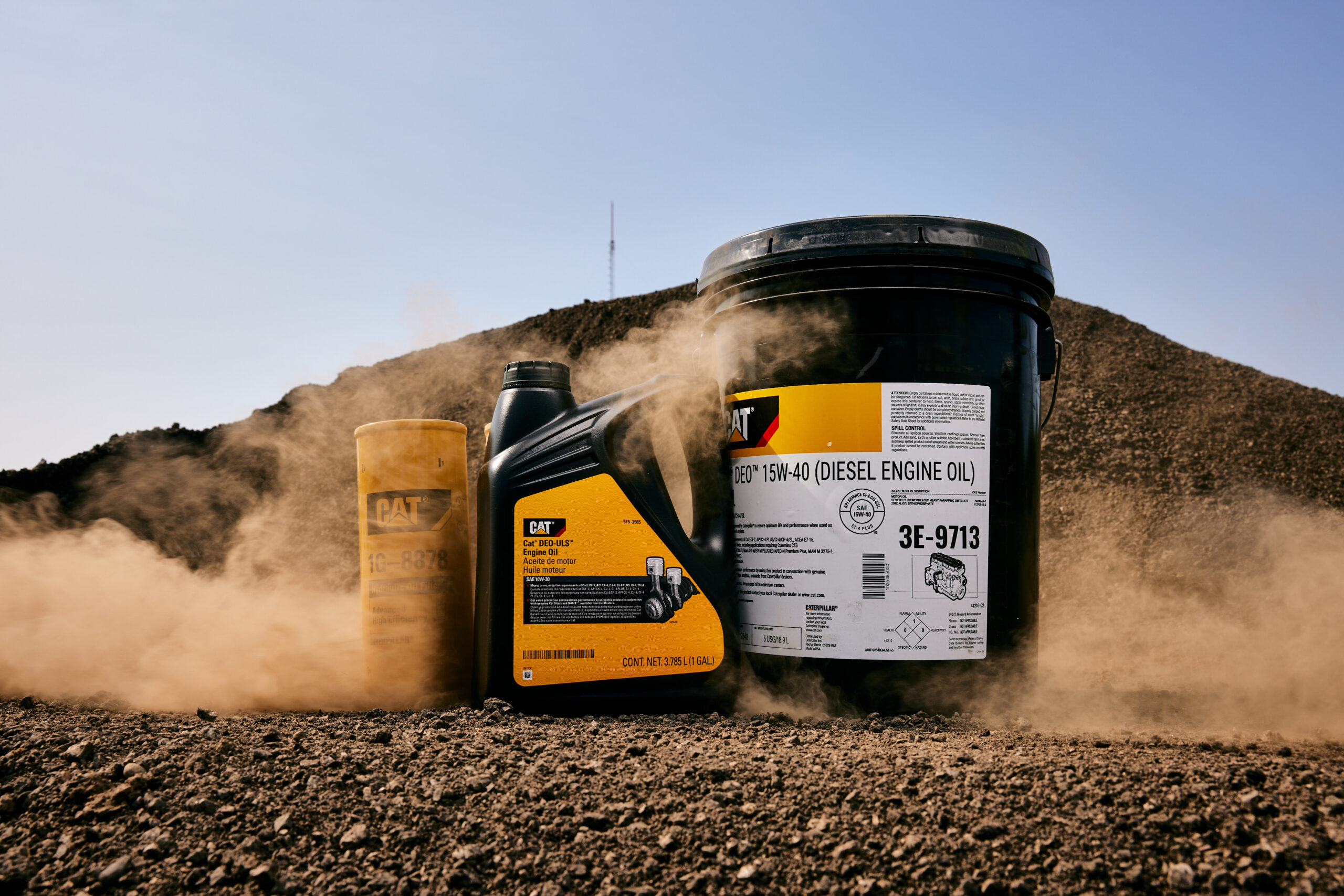 Cat 1G-8878 Hydraulic Oil filter, Engine oil gallon, and Diesel Engine oil tub posed in front of a mound of dirt and rocks. Some dust obstructs the parts